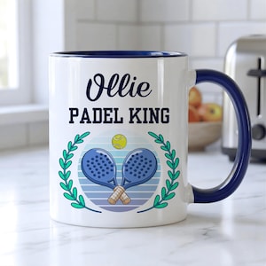Könnte beinhalten: Weißer Keramikbecher mit marineblauem Griff und Rand. Der Becher zeigt den Namen "Ollie" über den Worten "PADEL KING" in fetter Schrift. Eine Grafik von zwei blauen Padel-Schlägern und einem gelben Ball ist auf dem Becher zentriert, umgeben von einem grünen Lorbeerkranz.