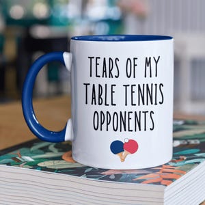 May include: White ceramic coffee mug with a blue rim and handle. The mug has black text that reads "Tears of my table tennis opponents" with a red and blue table tennis paddle graphic.