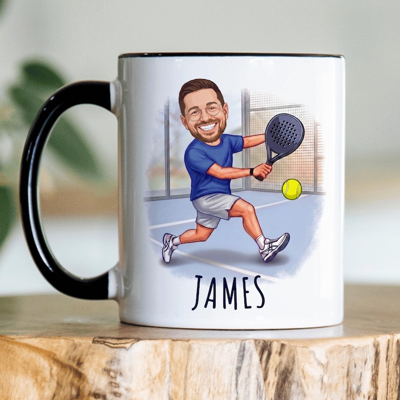 Puede incluir: Taza de cer&aacute;mica blanca con asa y borde negros. La taza presenta una ilustraci&oacute;n de dibujos animados de una persona jugando al p&aacute;del, con el nombre "JAMES" debajo. Tambi&eacute;n es visible una pelota de tenis amarilla.