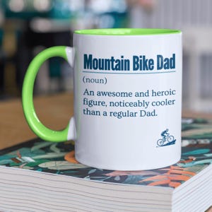 May include: White ceramic mug with a green rim and handle. The mug has a blue and white design that reads "Mountain Bike Dad (noun) An awesome and heroic figure, noticeably cooler than a regular Dad." A small illustration of a person riding a mountain bike is at the bottom of the mug.