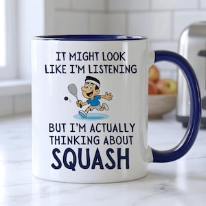 May include: White ceramic mug with a blue interior and handle. The mug features the text "IT MIGHT LOOK LIKE I'M LISTENING BUT I'M ACTUALLY THINKING ABOUT SQUASH" with a cartoon illustration of a squash player.
