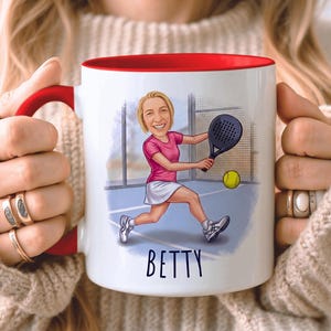 Puede incluir: Taza de cer&aacute;mica blanca con interior y asa rojos. La taza presenta una ilustraci&oacute;n de dibujos animados de una persona jugando al p&aacute;del, con el nombre "BETTY" en escritura negra. La persona lleva una camisa rosa y una falda blanca.