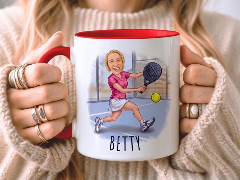 Puede incluir: Taza de cer&aacute;mica blanca con interior y asa rojos. La taza presenta una ilustraci&oacute;n de dibujos animados de una persona jugando al p&aacute;del, con el nombre "BETTY" en escritura negra. La persona lleva una camisa rosa y una falda blanca.