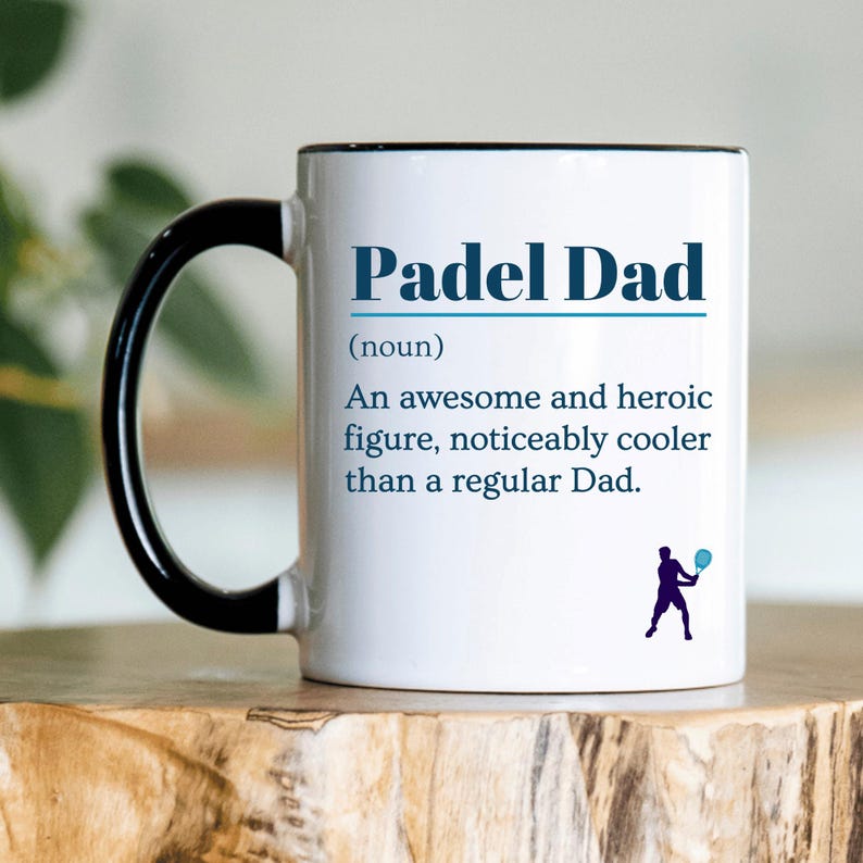 May include: White ceramic mug with a black handle and rim. The mug features the text "Padel Dad" in blue, along with a definition and a silhouette of a person playing padel. The mug is sitting on a wooden surface.