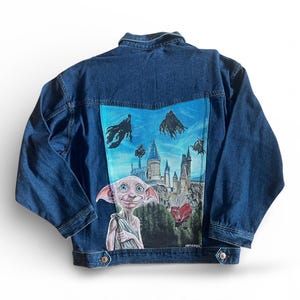 May include: A blue denim jacket featuring a painted design on the back. The artwork shows a castle, characters, and flying figures against a blue sky. The jacket has a button-down front and two chest pockets.
