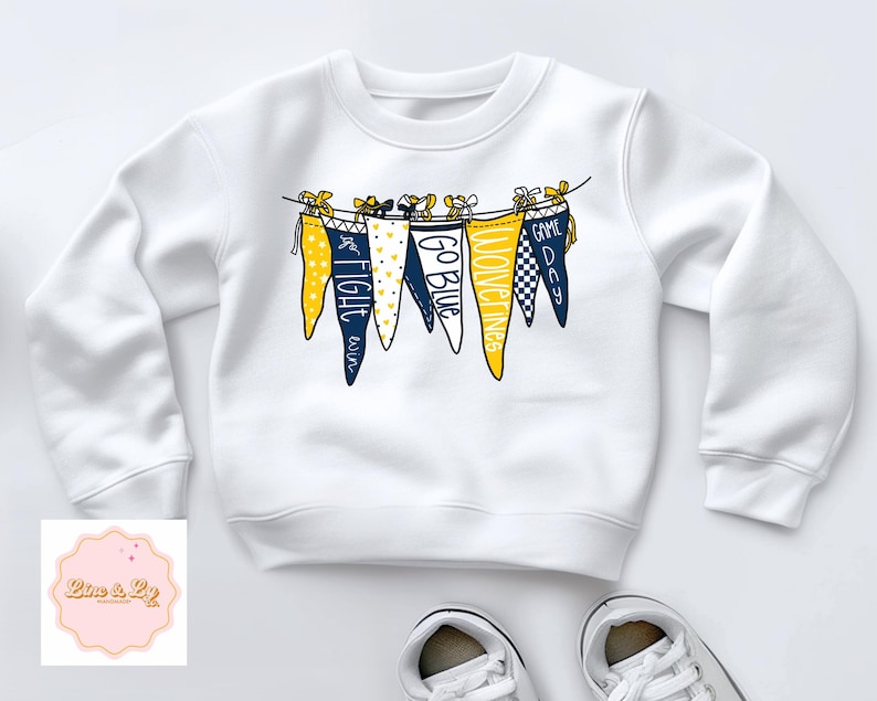 May include: White sweatshirt with a navy blue and yellow pennant banner design. The pennants have the words "Game Day", "Wolverines", "Go Blue", "Fight Blue", and "Fight  'em" printed on them.