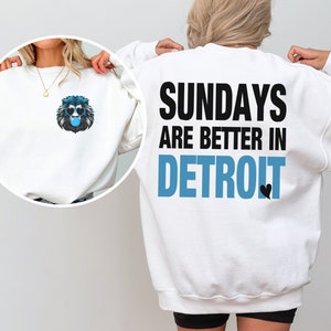 Puede incluir: Sudadera blanca con un gr&aacute;fico de le&oacute;n con gafas de sol azules y una burbuja de chicle en la parte delantera. La parte trasera de la sudadera tiene el texto "SUNDAYS ARE BETTER IN DETROIT" en negro y azul.