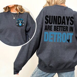 Puede incluir: Sudadera gris oscuro con el texto "SUNDAYS ARE BETTER IN DETROIT" en negro y azul. Un peque&ntilde;o gr&aacute;fico de un le&oacute;n con gafas de sol y una burbuja est&aacute; en la parte delantera. La sudadera tiene cuello redondo.