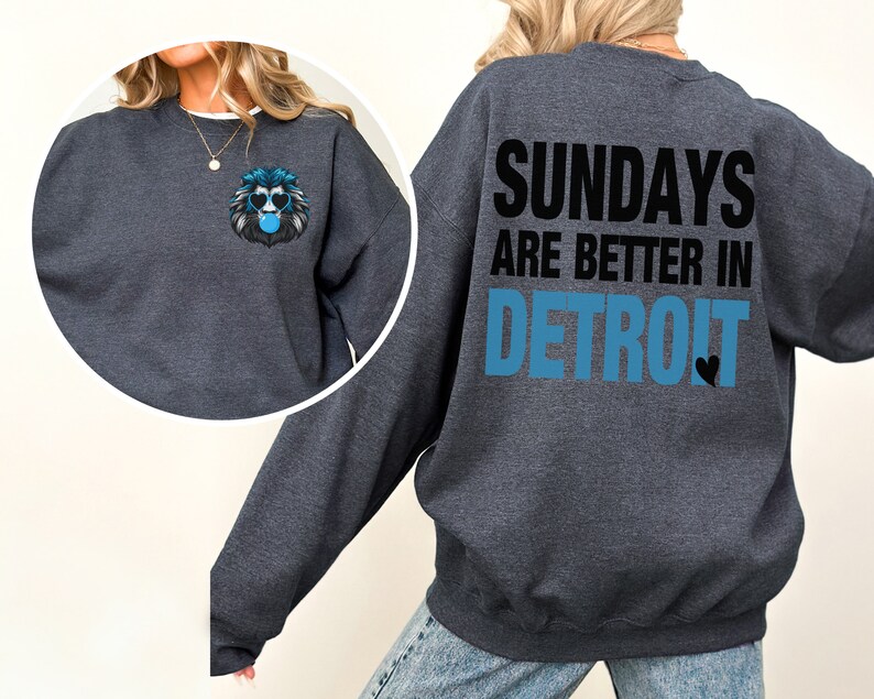 Puede incluir: Sudadera gris oscuro con el texto "SUNDAYS ARE BETTER IN DETROIT" en negro y azul. Un peque&ntilde;o gr&aacute;fico de un le&oacute;n con gafas de sol y una burbuja est&aacute; en la parte delantera. La sudadera tiene cuello redondo.