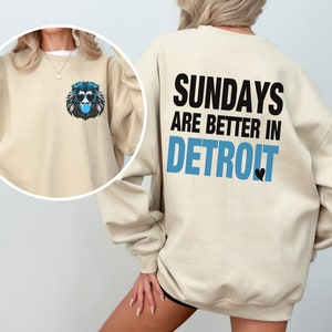 Puede incluir: Sudadera beige con un gr&aacute;fico de le&oacute;n con gafas de sol y soplando una burbuja en la parte delantera. La parte trasera de la sudadera tiene el texto "SUNDAYS ARE BETTER IN DETROIT" en negro y azul, con un peque&ntilde;o coraz&oacute;n negro.