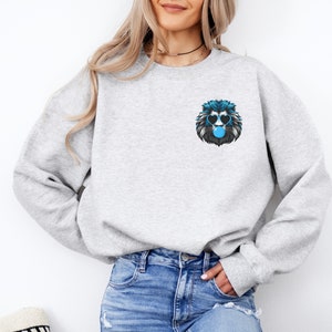 Puede incluir: Una sudadera gris claro con un gr&aacute;fico de le&oacute;n azul y negro en el pecho. El le&oacute;n lleva gafas de sol en forma de coraz&oacute;n y sopla una burbuja. La sudadera est&aacute; hecha de un material suave.