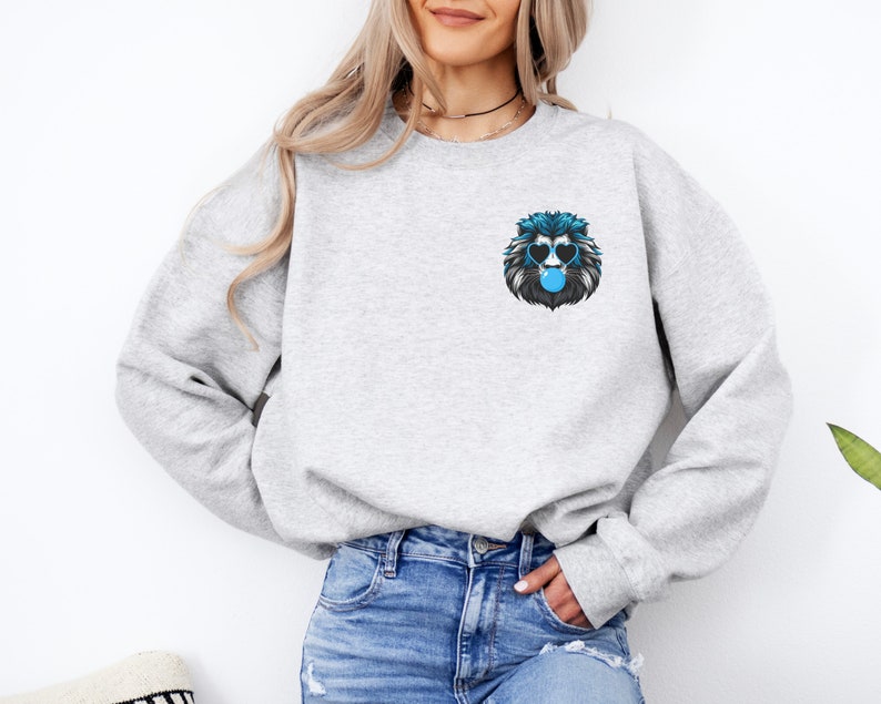 Puede incluir: Una sudadera gris claro con un gr&aacute;fico de le&oacute;n azul y negro en el pecho. El le&oacute;n lleva gafas de sol en forma de coraz&oacute;n y sopla una burbuja. La sudadera est&aacute; hecha de un material suave.