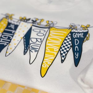 May include: White t-shirt with a graphic of pennant flags that say "Go Blue", "Wolverines", "Game Day", and "Fight" in yellow and blue lettering.
