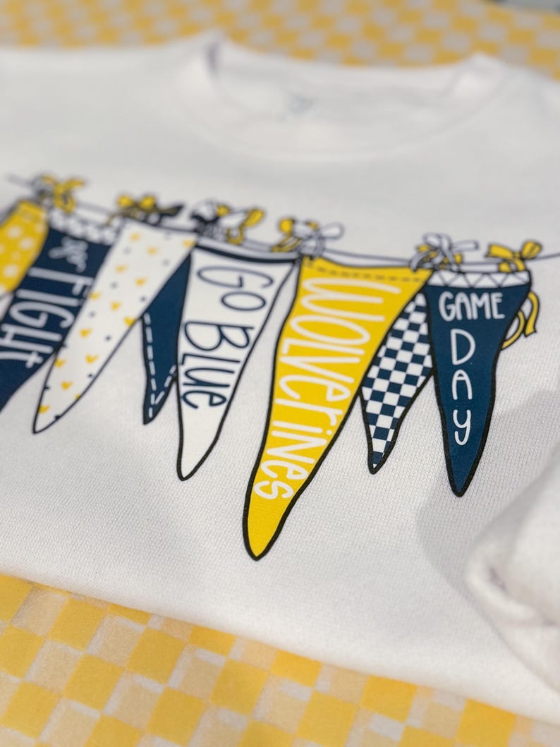 May include: White t-shirt with a graphic of pennant flags that say "Go Blue", "Wolverines", "Game Day", and "Fight" in yellow and blue lettering.