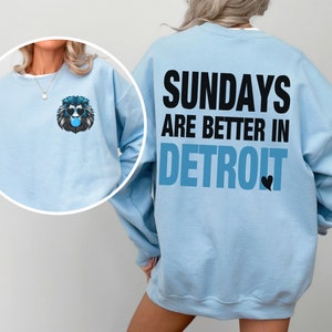 Puede incluir: Sudadera azul claro con el texto "SUNDAYS ARE BETTER IN DETROIT" en la espalda. Un gr&aacute;fico de le&oacute;n con gafas de sol y una burbuja de chicle est&aacute; en la parte delantera. Estilo casual.