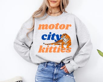 May include: A light gray sweatshirt with the words "motor city kitties" in orange and blue. A cartoon tiger in orange and black is centered below the word "city". The sweatshirt is made of a soft material.