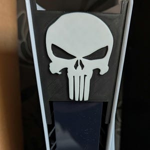 Puede incluir: Un logotipo de calavera blanca sobre un panel negro, diseñado para una consola de juegos. La calavera tiene una expresión amenazante con cuencas de los ojos detalladas y un diseño de líneas verticales debajo. El panel está montado en una consola blanca y negra.