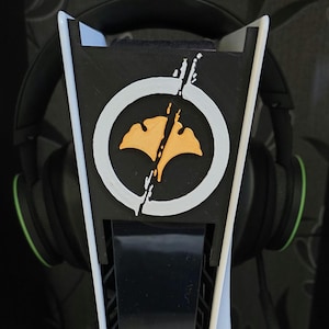 May include: A white and black gaming console stand with a circular design featuring a yellow leaf-like shape. A pair of black headphones with green accents rests on the stand. The stand is designed to hold the console.