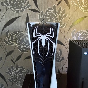 Puede incluir: Una consola PlayStation 5 blanca y negra con un diseño personalizado de Spider-Man. La consola tiene un diseño de telaraña negra con una araña blanca en el centro.