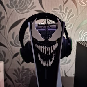 May include: A black and white 3D printed headphone stand, featuring a Venom symbiote design. The stand is made of plastic and has a spiderweb pattern on the back.