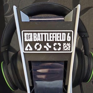 Może przedstawiać: Czarno-biały stojak na słuchawki z napisem "BF Battlefield 6" i symbolami. Czarny zestaw słuchawkowy z zielonymi akcentami spoczywa na stojaku. Akcesorium dla graczy.