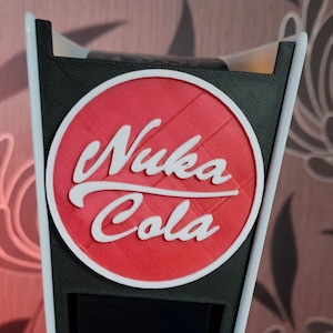 Puede incluir: Una funda personalizada para consola PlayStation 5 con el logotipo de Nuka Cola. La funda es roja con un logotipo y texto circular blanco. La consola es blanca y negra.