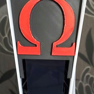 Puede incluir: Una consola de juegos blanca con una cubierta negra impresa en 3D que presenta una letra griega Omega roja.