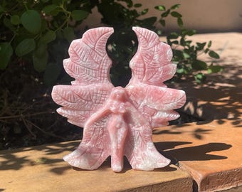 May include: A pink stone carving of an angel with outstretched wings. The angel's wings are detailed with carved feather patterns. The sculpture is set against a natural background, highlighting the pink and white hues of the stone.