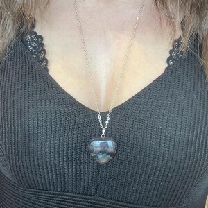Puede incluir: Collar de cadena plateada con un colgante en forma de coraz&oacute;n. El colgante es una piedra transl&uacute;cida y pulida con tonos morados y grises. El collar se lleva sobre un top negro.
