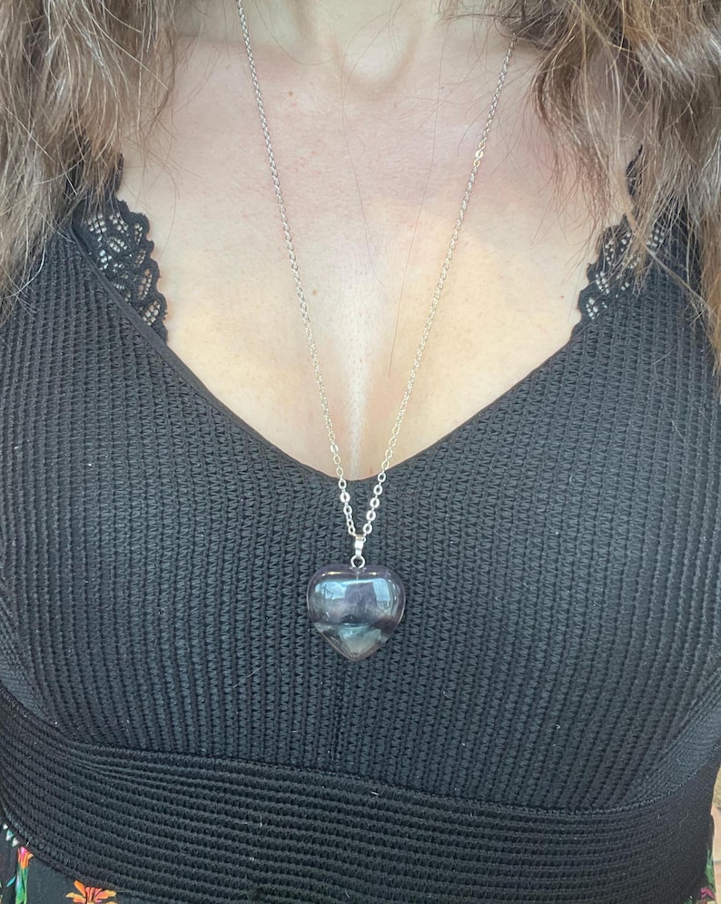 Puede incluir: Collar de cadena plateada con un colgante en forma de coraz&oacute;n. El colgante es una piedra transl&uacute;cida y pulida con tonos morados y grises. El collar se lleva sobre un top negro.