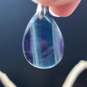 以下が含まれることがあります： 銀色のベイルが付いた、涙の形をしたペンダント。ペンダントは、紫、青、透明な色の層を持つ半透明の石でできています。石は銀色のベイルで保持されており、人の指で支えられています。