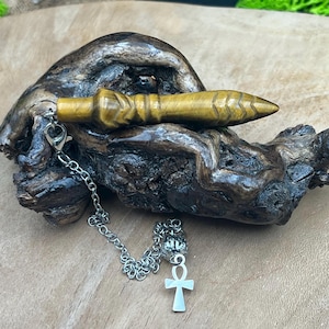 Puede incluir: Un péndulo amarillo dorado con punta y detalles tallados, conectado a una cadena plateada y un dije Ankh. El péndulo descansa sobre un trozo de madera oscura y texturizada.