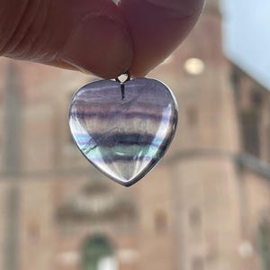Puede incluir: Un colgante en forma de coraz&oacute;n hecho de fluorita transl&uacute;cida e iridiscente. La piedra muestra bandas de color p&uacute;rpura, verde y blanco. El colgante est&aacute; suspendido de un peque&ntilde;o lazo de metal.