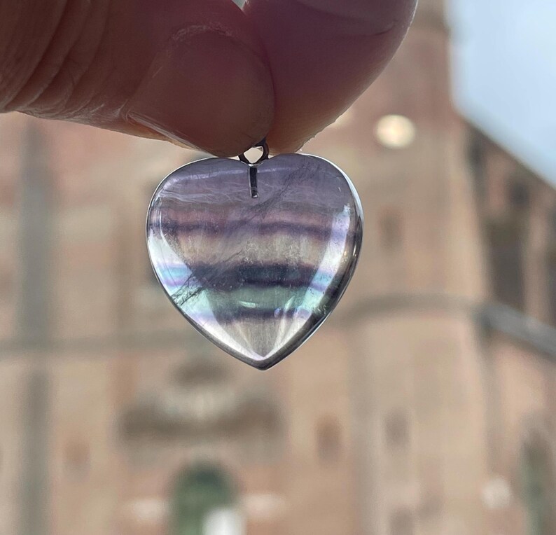 Puede incluir: Un colgante en forma de coraz&oacute;n hecho de fluorita transl&uacute;cida e iridiscente. La piedra muestra bandas de color p&uacute;rpura, verde y blanco. El colgante est&aacute; suspendido de un peque&ntilde;o lazo de metal.