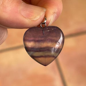 Puede incluir: Un colgante en forma de coraz&oacute;n de fluorita pulida. La piedra muestra bandas de tonos morados, lavanda y amarillo-marr&oacute;n. Una peque&ntilde;a fianza plateada est&aacute; unida en la parte superior para colgar. El colgante est&aacute; sostenido por los dedos de una persona.