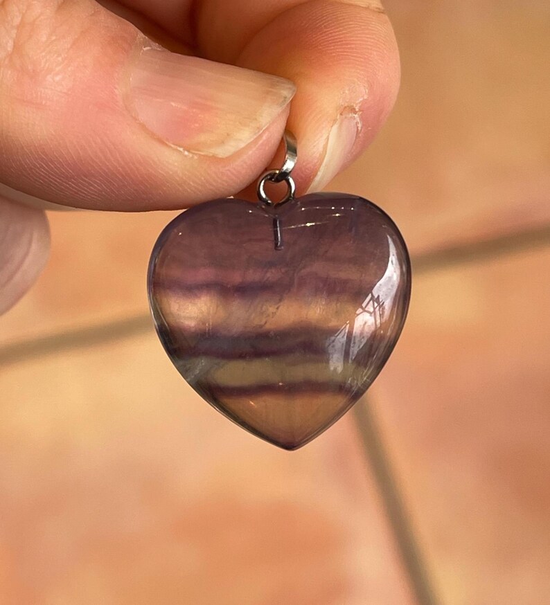 Puede incluir: Un colgante en forma de coraz&oacute;n de fluorita pulida. La piedra muestra bandas de tonos morados, lavanda y amarillo-marr&oacute;n. Una peque&ntilde;a fianza plateada est&aacute; unida en la parte superior para colgar. El colgante est&aacute; sostenido por los dedos de una persona.