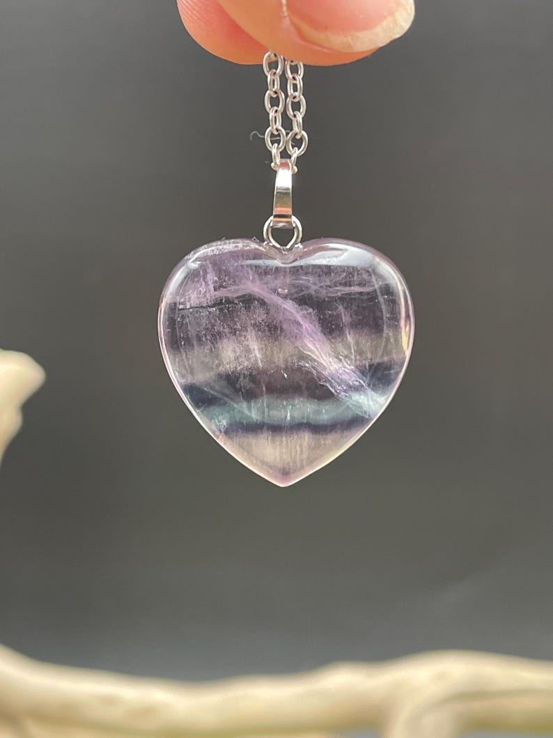 Puede incluir: Colgante en forma de coraz&oacute;n hecho de piedra transl&uacute;cida, con tonos de morado, blanco y azul. Cuelga de una delicada cadena de plata. Los patrones naturales de la piedra crean un dise&ntilde;o &uacute;nico y llamativo. Una joya.