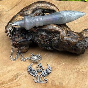 Op de afbeelding: Een grijze labradoriet pendel met een gladde, taps toelopende vorm en een zilverkleurige metalen ketting. De ketting is voorzien van een kleine, getextureerde bolvormige kraal en een platte, zilverkleurige metalen bedel van een gevleugeld katachtig figuur.