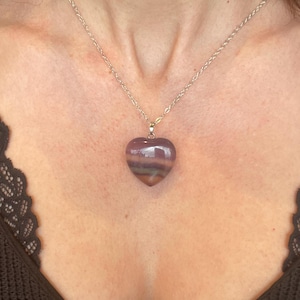 Puede incluir: Un collar con colgante en forma de coraz&oacute;n. El colgante es de piedra morada y verde. La cadena del collar es de color plateado. El colgante cuelga de la cadena y descansa sobre el pecho.