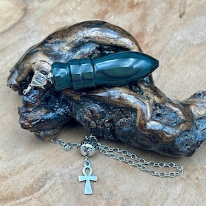 Puede incluir: Un péndulo verde oscuro con una cadena plateada y un dije de Ankh. El péndulo es cilíndrico con un extremo puntiagudo. La cadena está unida a una pequeña cuenta plateada y un cierre plateado decorativo. El péndulo descansa sobre un trozo de madera oscura.