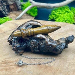 Peut inclure: Un pendule en bois avec une pointe et des détails sculptés, relié à une chaîne argentée et une petite boule texturée. Le pendule repose sur un morceau de bois foncé et texturé.