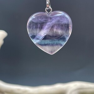 Puede incluir: Collar con colgante en forma de coraz&oacute;n y cadena plateada. El colgante est&aacute; hecho de una piedra transl&uacute;cida, morada y azul. La piedra tiene un aspecto en capas, con vetas de color. El collar se muestra sobre un fondo borroso.