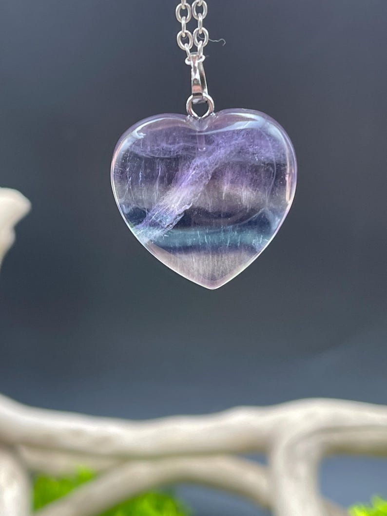 Puede incluir: Collar con colgante en forma de coraz&oacute;n y cadena plateada. El colgante est&aacute; hecho de una piedra transl&uacute;cida, morada y azul. La piedra tiene un aspecto en capas, con vetas de color. El collar se muestra sobre un fondo borroso.
