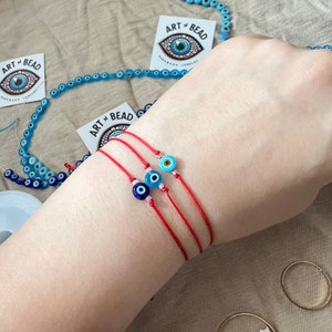 May include: Three red string bracelets with blue and white evil eye charms. The bracelets are on a person's wrist. The image also shows a string of blue and white evil eye beads and two gold rings.