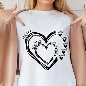 Puede incluir: Camiseta blanca con un diseño de corazón negro. El corazón está formado por varios corazones superpuestos. El texto "ADD TEXT" aparece dos veces en el corazón. El texto "NAME" aparece varias veces alrededor del corazón.