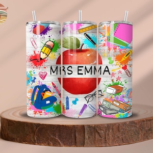 May include: Three stainless steel tumblers with colourful designs featuring a red apple, pencils, and other school supplies. The tumblers have a white background with black text that reads "Mrs. Emma".