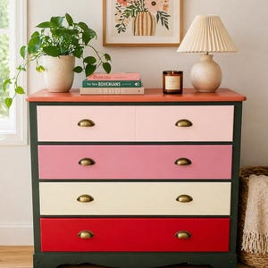 May include: A colorful dresser with drawers in shades of pink, cream, and red, accented with brass hardware. The top is coral, and the base is dark green. A lamp, books, and a candle sit on top, with a framed floral print in the background.