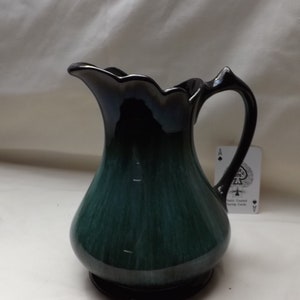 May include: A dark green and black ceramic jug with a distinctive, scalloped rim and a curved handle. The jug has a rounded base and a glossy finish, suitable for serving drinks. The design is reminiscent of vintage or antique styles.