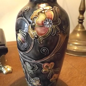 May include: A dark grey ceramic vase featuring a floral pattern. The flowers are in shades of yellow, orange, and red, with green leaves and stems. The vase has a rounded shape and a narrow neck. The design is outlined in a lighter colour.