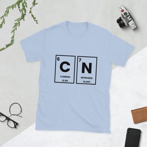 May include: A light blue t-shirt featuring the chemical symbols for carbon and nitrogen, with the atomic numbers and weights displayed. The design is in black and white, creating a minimalist and scientific aesthetic.